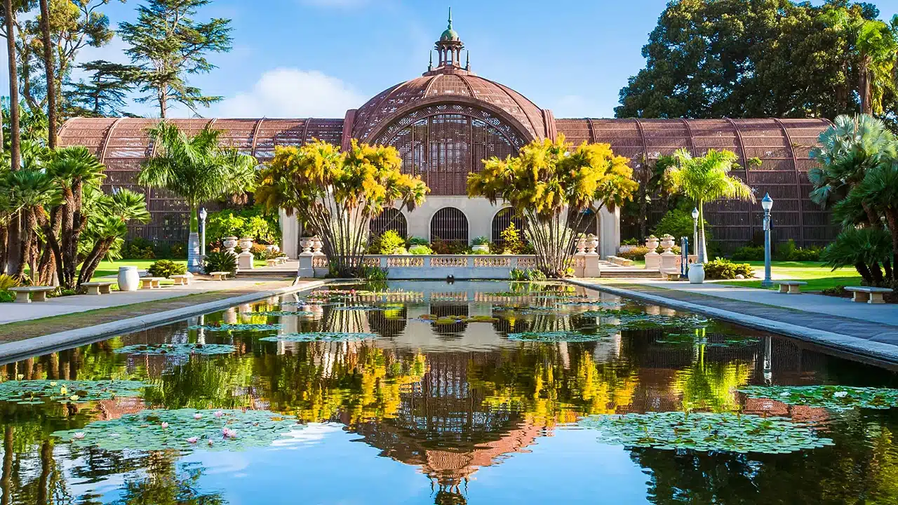 A large domed greenhouse with arched windows is reflected in a pond with lily pads, surrounded by palm trees and landscaped gardens—a must-see when you visit San Diego.