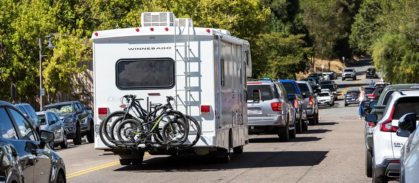 A white RV with bicycles mounted on back drives down a sunny, tree-lined street with traffic.