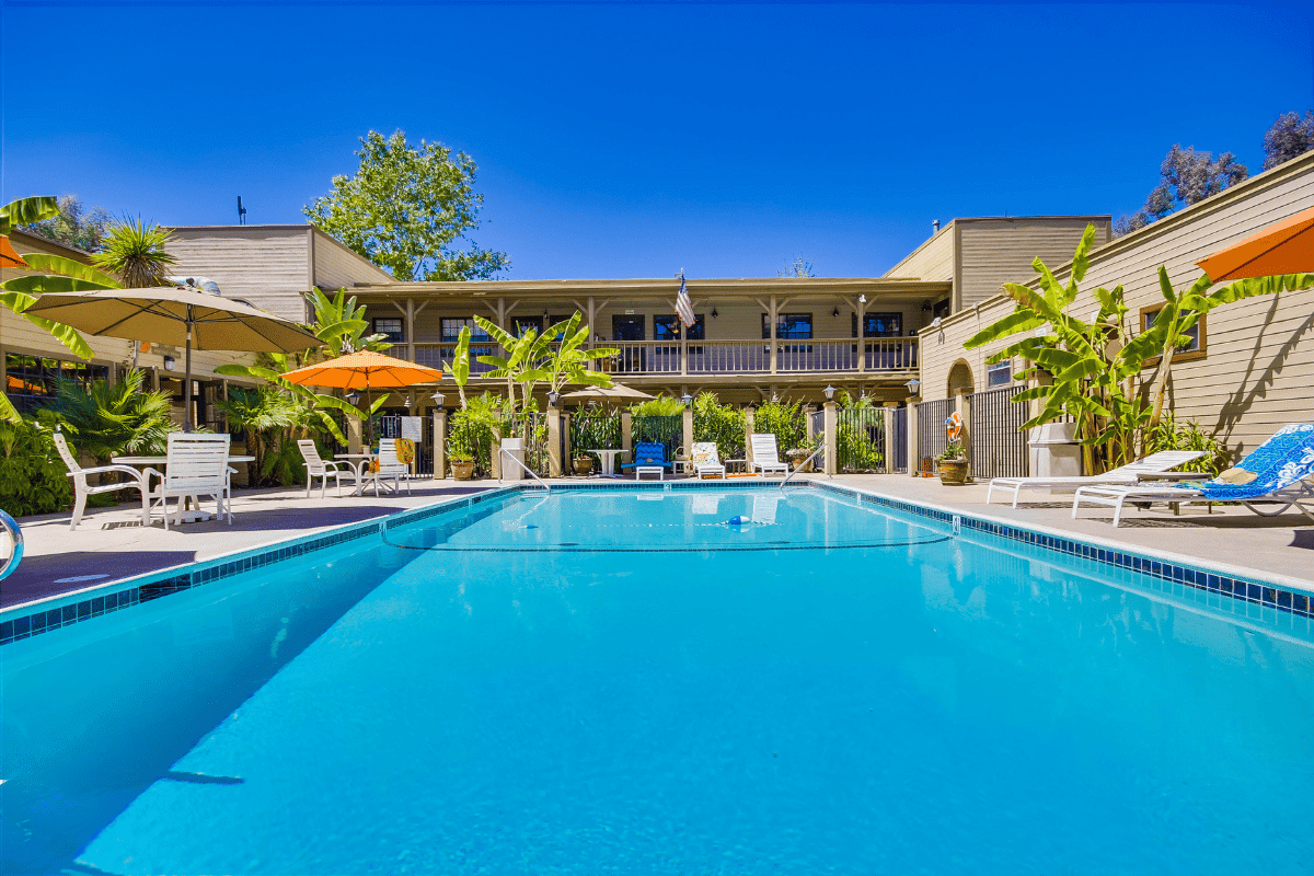 A rectangular outdoor swimming pool at Circle RV Resort is surrounded by lounge chairs, tables with umbrellas, and tropical plants, with a two-story building in the background.