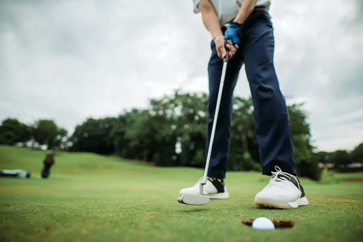 Person in golf attire poised to putt a golf ball on a green, with another person and trees visible in the background.