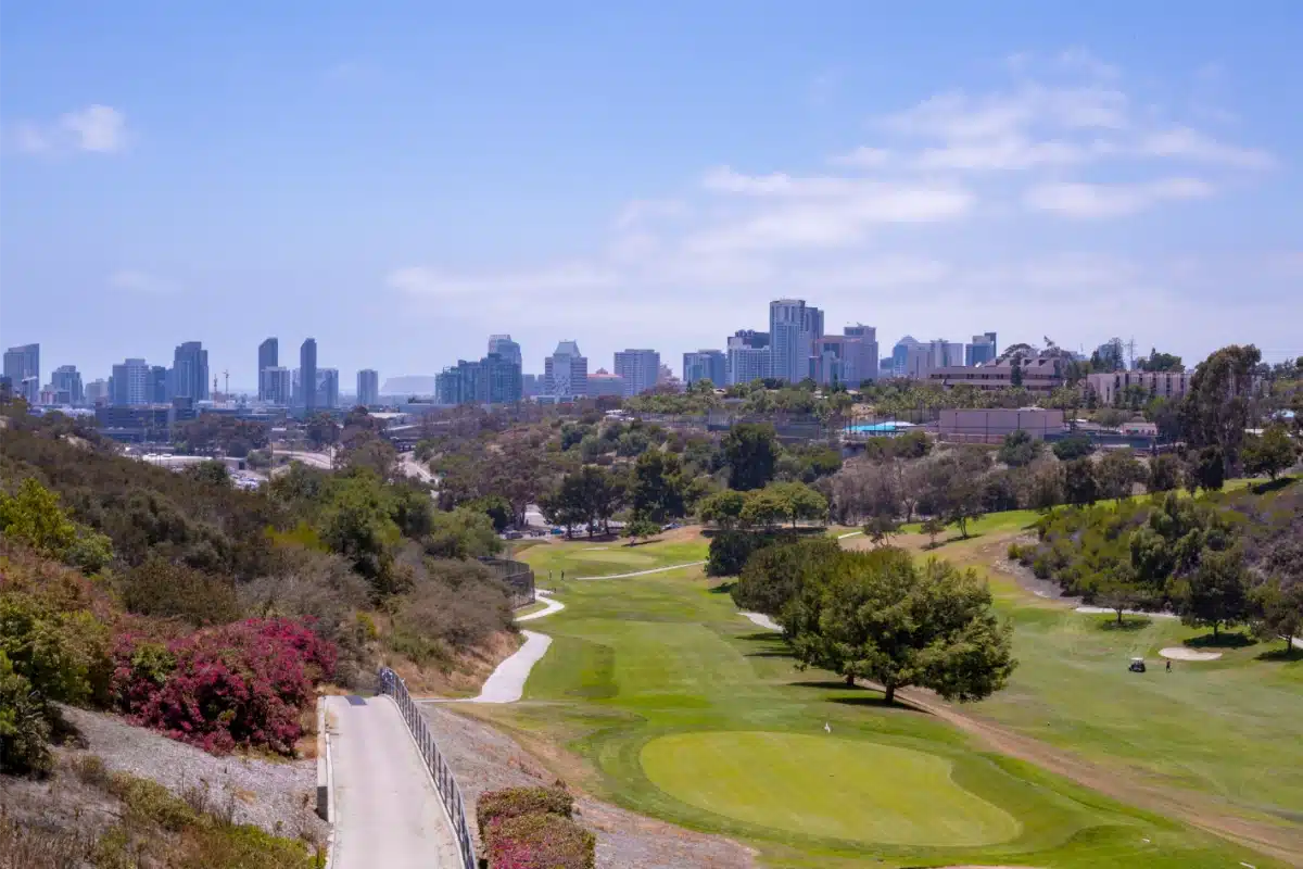 A golf course with green fairways and trees in the foreground, city skyline with tall buildings in the background under a partly cloudy sky.
