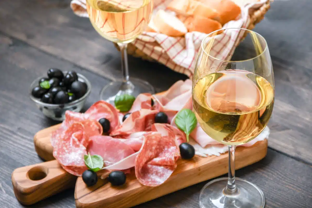 A wooden board with assorted cured meats, black olives, and basil leaves next to two glasses of white wine; a basket of bread is in the background—perfect for those who visit San Diego and enjoy local flavors.